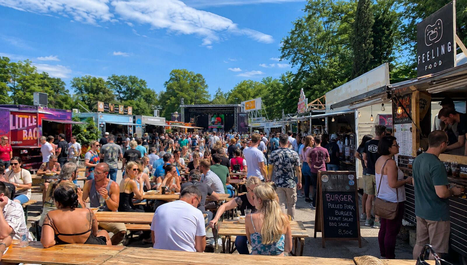 Besucherinnen und Besucher an Biertischgarnituren zwischen Foodtrucks bei einem sonnigen Street-Food-Festival im Park, im Hintergrund eine Bühne